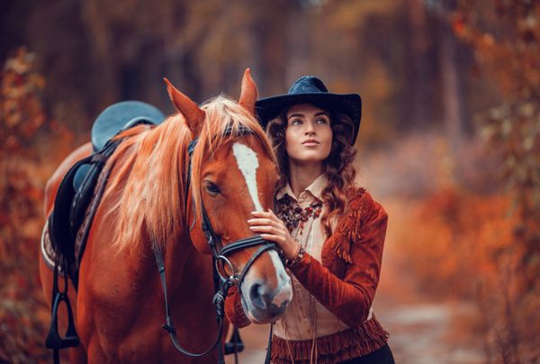 زیبا دختر شیک لباس پوشیدن به عنوان گاوچران آمریکایی در یک جاده روستایی با اسب saddled پاییز طلایی