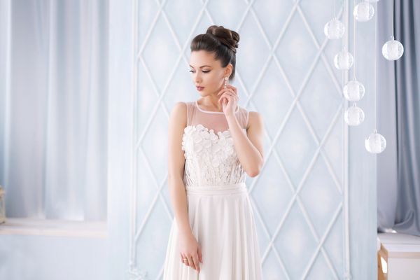 بانوی زیبا جوان در لباس عروسی سفید با دسته گل زدن در منزل لوکس ایستاده و به دنبال پایین پرتره