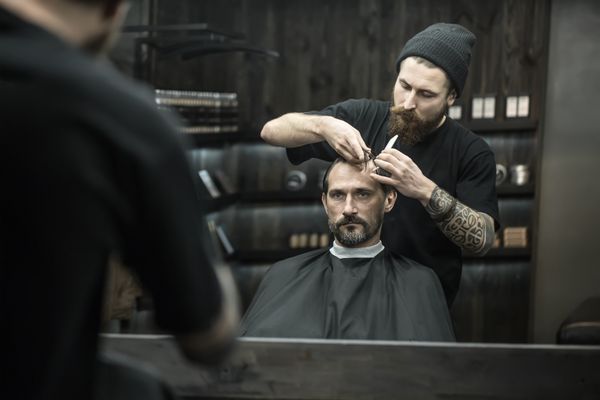 مرد ریشدار شیک در یک کلاه سیاه مو برش مو در آرایشگاه آرایشگری وحشیانه با یک کلاه سیاه با موهای خود را شانه می کند افقی