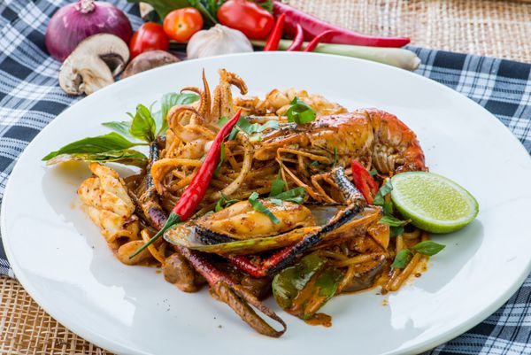 اسپاگتی سرخ شده تند با غذاهای دریایی و ریحان در ظرف سفیدی با قاشق و چنگال غذای همجوشی تایلندی با اسپاگتی بر روی میز چوبی سبک تایلندی