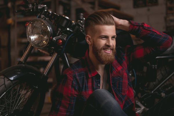 مرد خوش تیپ ریشو در لباس های گاه به گاه لبخند می زند در هنگام نشستن در موتور سیکلت در مغازه تعمیر