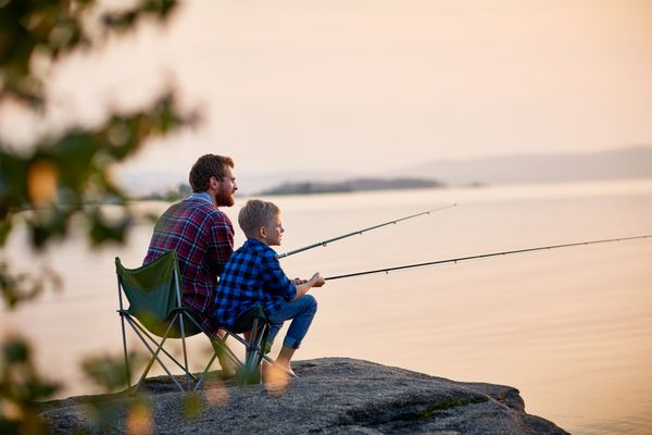 منظره جانبی تصویری از پدر و پسر که در کنار دریاچه های ماهیگیری با میله ها در کنار دریاچه آرام با منظره ای از خورشید قرار گرفته اند هم پوشیدن پیراهن های شطرنجی از پشت درخت شلیک می کنند