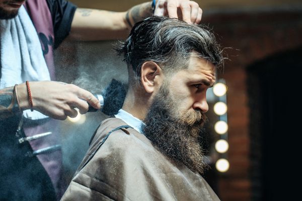 مرد ریش دار با ریش بلند وحشیانه قهوه ای قهوه ای با سبیل با موهای شیک مو گرفتن پودر روی پوست با استفاده از برس آرایش توسط سلمانی یا دست های آرایشگری در آرایشگاه