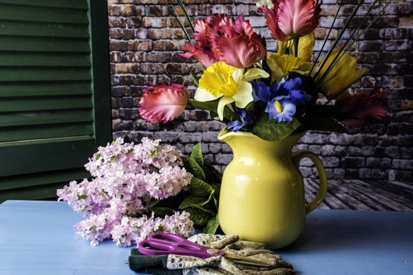 دسته گل تازه بهار گل گلدان آب زرد در جدول آبی با کلیپرز و دستکش کار با شاتر سبز و پس زمینه آجری دیوار