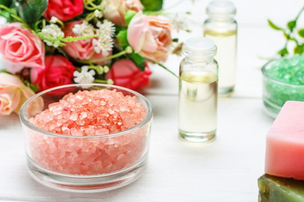 نمک دریای سرخ دو بطری با روغن های آرایشی و گل رز در یک میز چوبی سفید