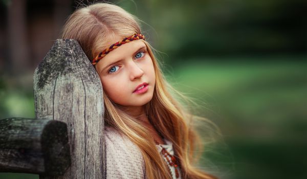 پرتره یک دختر زیبا با چشم آبی در یک لباس پرنعمت سفید و مد لوازم جانبی ایستاده در نزدیکی حصار چوبی مدل جوان نشان دادن در سبک بوهمی کودک بسیار به دوربین نگاه می کند