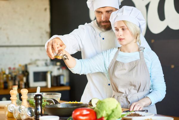 سرآشپز میان ساله متمرکز و زن جوان بسیار زیبا که در حال پر کردن روغن زیتون به همراه سرپا نگه داشتن ظرف در کارگاه آشپزی