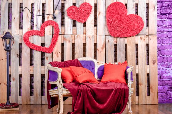 The decor of the interior is an old vintage sofa of ultra violet colored wood with red pillows and decorated hearts on the background of wooden pallets Valentine
