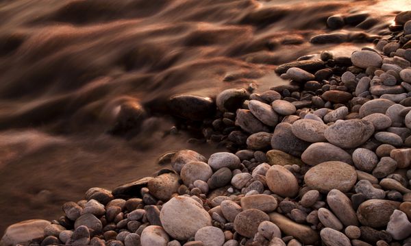 یک تصویر الهام بخش آرام از یک رودخانه که در نزدیکی بستر سنگی که توسط نور غروب خورشید روشن می شود جریان دارد