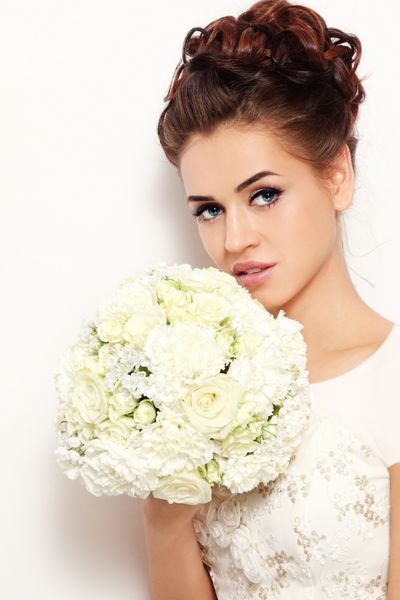 عروس زیبا با آرایش شیک و دسته گل نگه داشتن مدل مو در دست خود بالای دیوار سفید