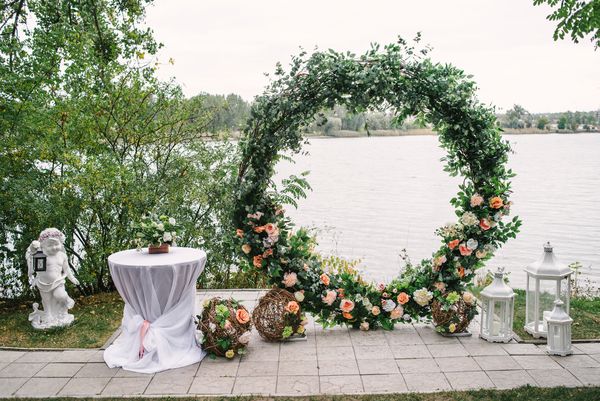 قوس عروسی گرد زیبا که با گل و فضای سبز در نزدیکی دریاچه یا رودخانه در فضای باز تزئین شده است فضای کپی را تکمیل می کند تزئینات مراسم عروسی در هوای آزاد