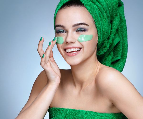 دختر خنده ای که از کرم مرطوب کننده روی صورت استفاده می کند عکس دختر جوان با پوست بی عیب و نقص در حوله های سبز مفهوم مراقبت از پوست و زیبایی