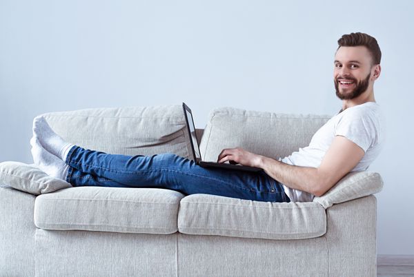 مرد جوان خوش تیپ ریشو در لباس های گاه به گاه کار با لپ تاپ در دست دراز کشیده بر روی مبل و نگاه کردن به دوربین نمای کنار آزاد کار در خانه