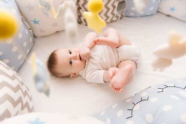 کودک ناز در یک تخت دور سفید قرار دارد مهد کودک سبک برای کودکان خردسال اسباب بازی های مخصوص نوزادان کودک لبخند را با موبایل از احساس و جویدن پا در اتاق خواب آفتابی
