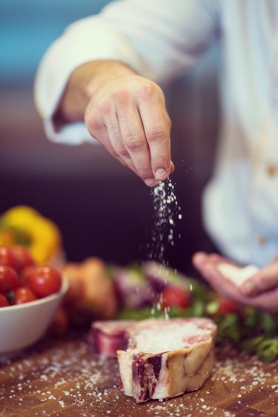 استاد سرآشپز با قرار دادن نمک برش آبدار استیک خام به همراه سبزیجات در یک میز چوبی قرار دارد
