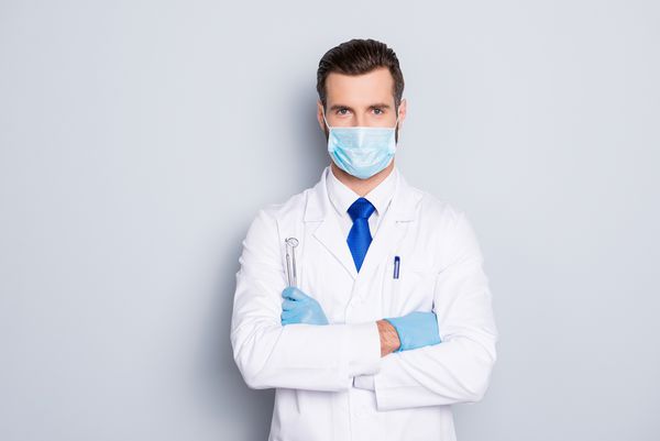 پرتره دندانپزشک خوش تیپ جذاب با مدل مو در ماسک محافظ صورت کت آزمایشگاه سفید کراوات آبی نگه داشتن تجهیزات در بازوها جدا شده در زمینه خاکستری