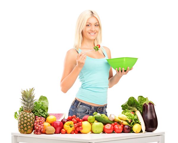 زن جوانی که سالاد می خورد پشت میز پر از میوه و سبزیجات ایستاده است