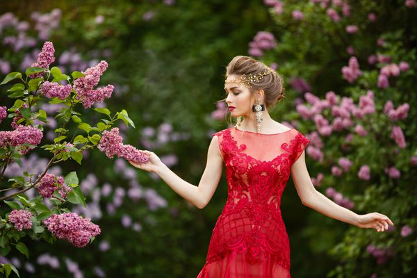 زن زرق و برق دار زیبا با لباس قرمز قرمز در باغ گل یاس شکوفه پرتره رشد در نور غروب خورشید دختر جوان جوان قفقازی در باغ قدم می زند و با لباس خود بازی می کند زمان بهار ملکه جوان