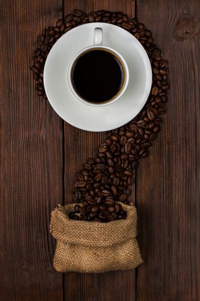 دانه های قهوه بو داده شده را از کیسه کوچک اطراف فنجان قهوه با نوشیدنی روی سطح چوبی تیره پراکنده می کند نمای بالا
