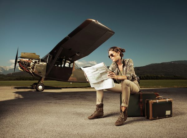 زن جوان زیبا که روی یک چمدان نشسته است به یک نقشه نگاه می کند هواپیما در پس زمینه