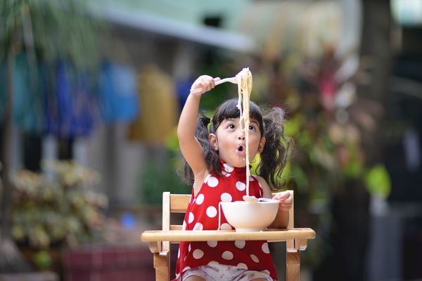دختر بچه ناز آسیایی هنگام خوردن سس سفید اسپاگتی ایتالیایی یا کربنارا روی میز در فضای باز سرگرم کننده است بچه ها با خوردن غذا سرگرم کننده و شاد هستند