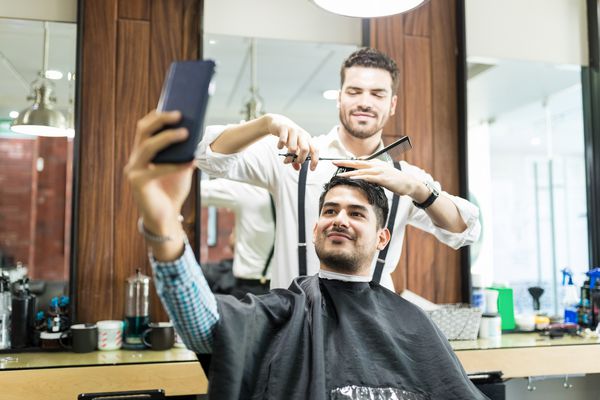 مشتری خوش تیپ و خوش سلیقه در حالی که آرایشگر موهای خود را در سالن می کند از تلفن همراه خود سلفی می کند