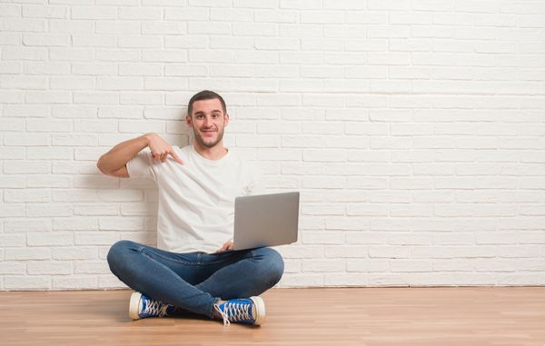 مرد جوان قفقازی که با استفاده از لپ تاپ رایانه ای با چهره غافلگیرانه انگشت خود را به سمت دیوار آجری سفید نشسته است