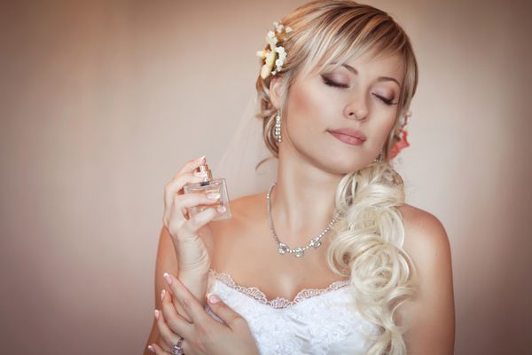 دختر عروس زیبا و خوشگل در لباس عروسی سفید با مدل مو و آرایش روشن و بطری عطر بانوی رمانتیک در لباس عروس و گل های مو آماده سازی نهایی برای عروسی است