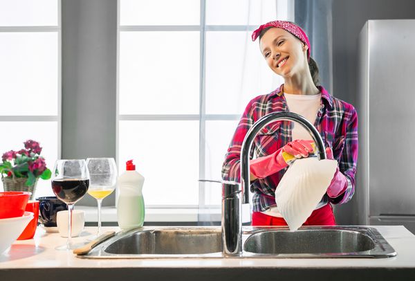 زن شاد در دستکشهای لاستیکی محافظ صورتی صفحه را در آشپزخانه شستشو می دهد