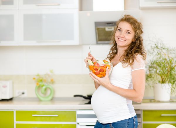 زن باردار در آشپزخانه که سالاد را میخورد لبخند می زندبه دوربین نگاه می کند