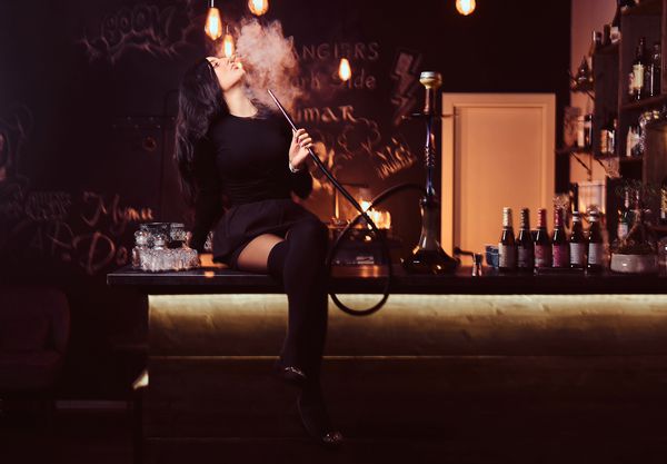 دختری با لباس سیاه اغواکننده قلیان را در حالی که در یک کلاب شبانه یا نوار بر روی میز نشسته است قلیان می کشید