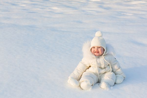 کودک زیبا در یک کت و شلوار برفی سفید که در یک روز آفتابی زمستانی روی برف تازه نشسته است