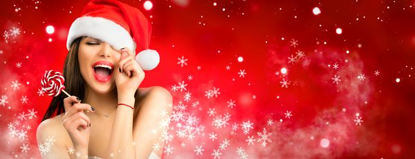 زن کریسمس دختر مدل زیبایی در کلاه بابا نوئل با لب های قرمز و آب نبات شیرین ابریشمی در دست خود شادی ابراز تعجب پرتره نزدیک روی زمستان برف زمستان قرمز گسترده با برف