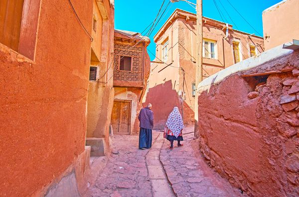 این زوج ارشد با لباس سنتی ابیاناکی در امتداد صعود باریک در میان خانه های خشتی قرمز ابیانه ایران قدم می زنند