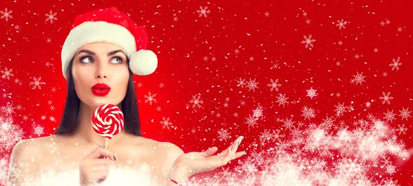 زن کریسمس دختر مدل زیبایی در کلاه بابا نوئل با لب های قرمز و آب نبات آب نبات چوبی lollipop با اشاره ارائه محصول حراجی ابراز تعجب پرتره closeup که در پس زمینه سفید جدا شده است