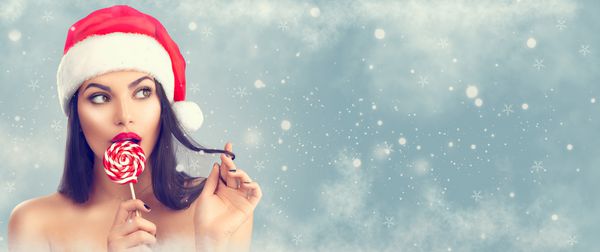زن کریسمس دختر مدل زیبایی در کلاه بابا نوئل با لب های قرمز و تفکر آب نبات شیرین ابریشمی کریسمس انتخاب هدیه پیشنهاد محصول بیان غافلگیر شده پرتره نزدیک در زمینه آبی