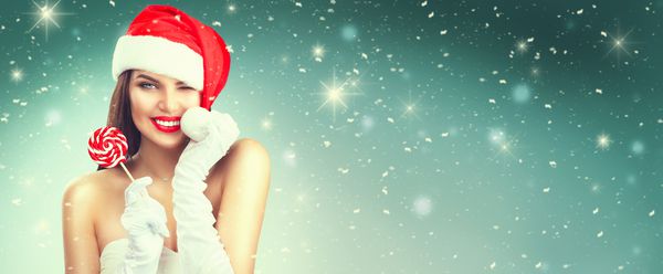 دختر کریسمس زن مدل خنده دار زیبایی در کلاه بابا نوئل با لب های قرمز دندان های سفید و آب نبات گل شیرین کریسمس با خنده حراجی ابراز تعجب پرتره نزدیک در زمینه قرمز