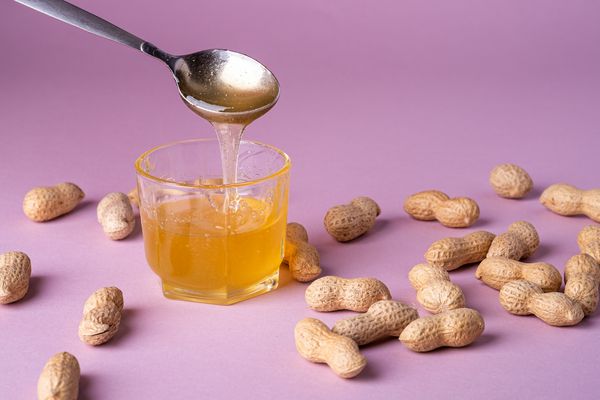 مایع عسل در لیوان با قاشق فلزی با بادام زمینی حداقل در زمینه زمینه بنفش فضای کپی بافتی مهره مواد غذایی