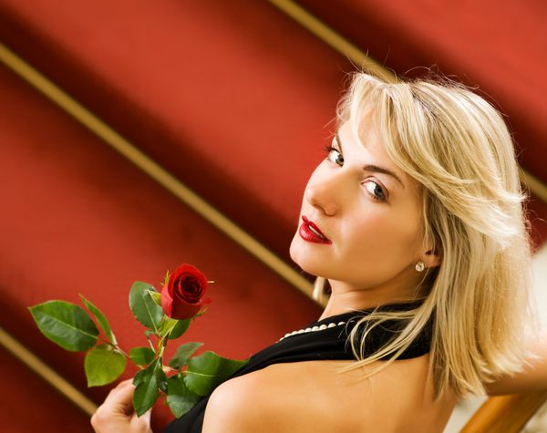 زن جوان زیبا که روی فرش قرمز ایستاده و گل رز را در دست دارد