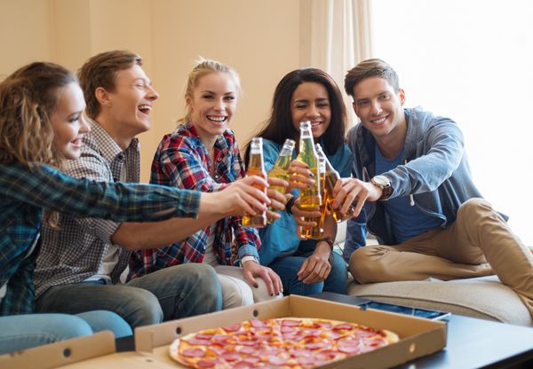 گروهی از دوستان جوان با پیتزا و بطری های نوشیدنی که در فضای داخلی خانه جشن می گیرند