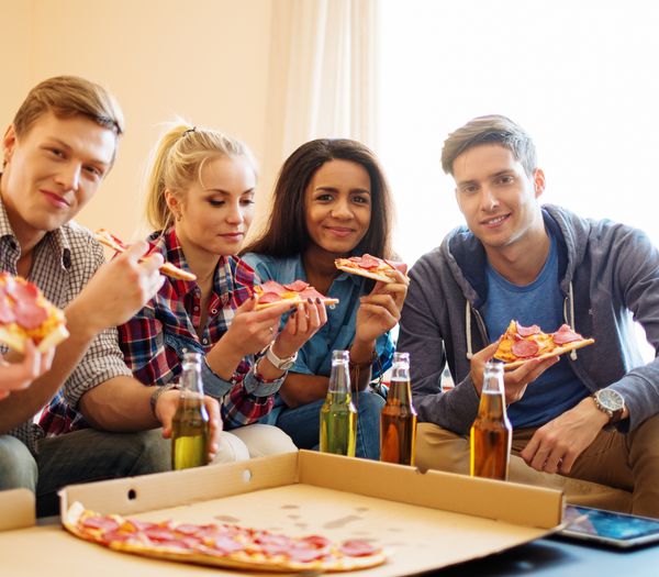 گروهی از دوستان چند قومی جوان با پیتزا و بطری های نوشیدنی که در فضای داخلی خانه جشن می گیرند