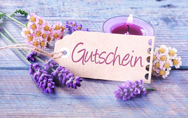 تگ هدیه با کلمه Gutschein به زبان آلمانی روی تخته های چوبی آبی روشن با شمع سوزان و اسطوخودوس و گل های تازه در یک مفهوم آبگرم و سلامتی