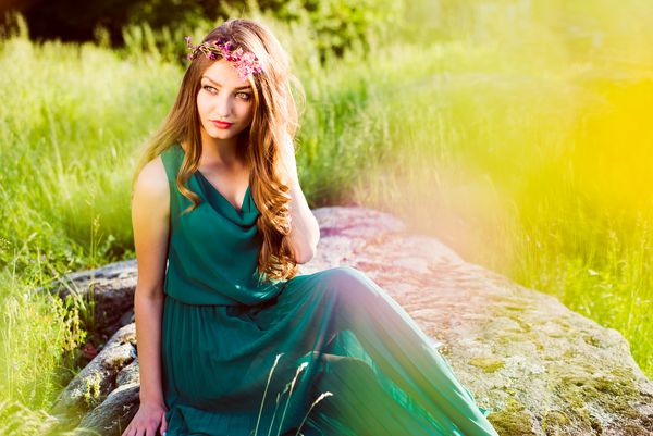پری زیبا دختر جوان زن ناز که بر روی سنگ با لباس سبز و تاج گل از گل amp amp؛ شعله های نور خورشید از پرتوهای amp amp؛ نگاه کردن به دوربین در پس زمینه فضای کپی تابستانی