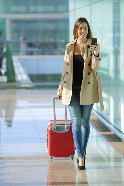 نمای جلوی یک زن مسافر که در راهرو فرودگاه در حال راه رفتن و استفاده از تلفن هوشمند است