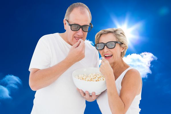 زن و شوهر بالغی که عینک سه بعدی دارند در حال خوردن پاپ کورن در برابر آسمان آبی روشن و ابرها هستند