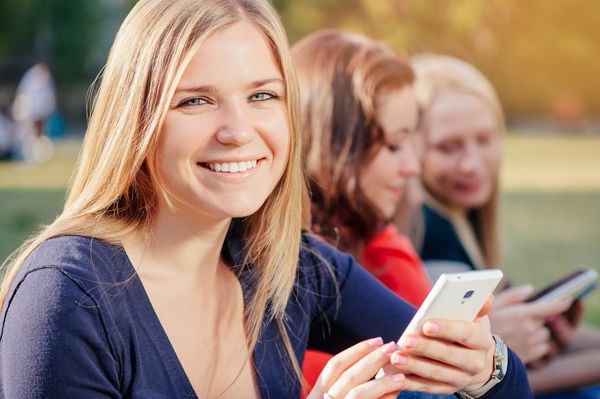 به اشتراک گذاری خبرهای خوب با دوستان زن جوان شاد که یک تلفن همراه داشت و لبخند می زد در حالی که گروهی از افراد بدون ارتباط از پس زمینه در حال برقراری ارتباط بودند