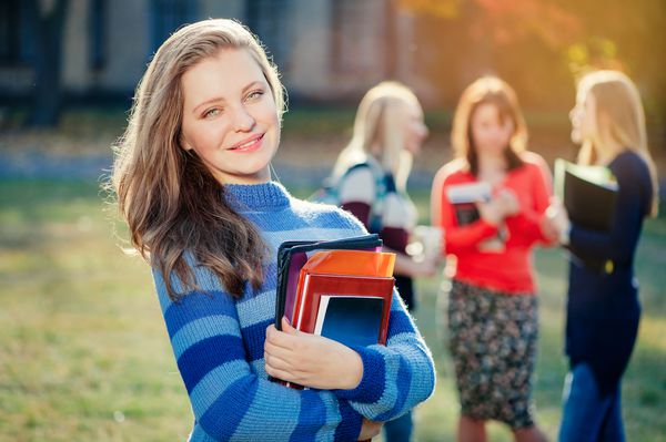 آینده من در دست من است زن جوان قفقازی جوانی که در حال ایستادن در نزدیکی ساختمان دانشگاه و همراه دوستانش است که در حال نگه داشتن کتاب است و لبخند می زند