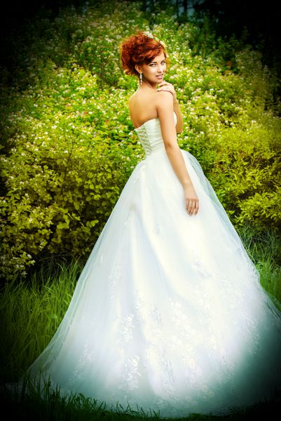 عروس با موهای قرمز زیبا و زیبا روی زمین چمن ایستاده است لباس عروسی و لوازم جانبی