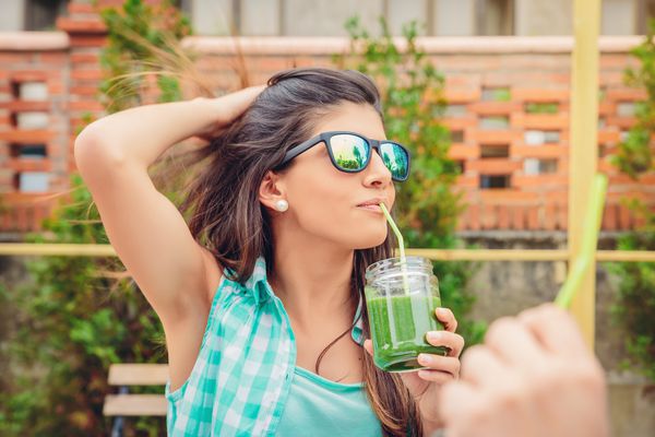 زن جوان و زیبا با عینک آفتابی که نوشابه سبزیجات سبز را با نی در روز تابستان در فضای بیرون می نوشد مفهوم نوشیدنی های ارگانیک سالم