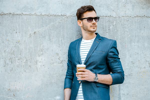 پرتره مرد جوان خوش تیپ و شیک با موهای ایستاده در فضای باز و تکیه دادن به دیوار مردی که ژاکت عینک آفتابی پیراهن و فنجان قهوه را در دست گرفته است